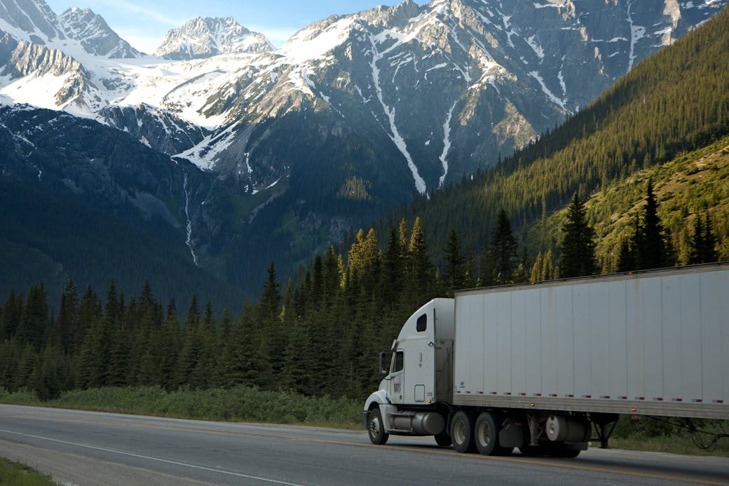 A semi-truck travels along a highway with snow-capped mountains in the background.